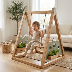 Puede incluir: Un columpio infantil de madera con estructura en forma de A, acabado en madera clara natural, cuerdas gruesas y paneles laterales en color verde salvia decorados con siluetas de montañas y jardineras integradas con plantas verdes.