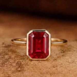 May include: A gold ring featuring a rectangular emerald-cut red gemstone set in a smooth bezel setting. The band is thin and polished, showcasing a clean design that highlights the vibrant red color and faceted edges of the central stone.