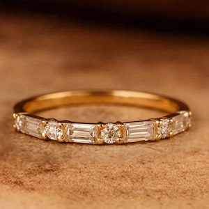 May include: A gold band ring featuring an alternating pattern of clear baguette-cut and round-cut stones set along the top half of the band. The metal has a polished yellow gold finish, and the stones are arranged in a repeating sequence of baguette and round.
