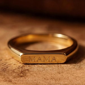 May include: A gold-toned ring featuring a flat, rectangular front surface with the word MAMA engraved in capital letters. The band has a smooth, polished finish and a consistent thickness throughout its circular design, showcasing a metallic lustre.