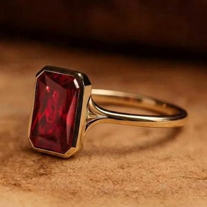 Peut inclure: Une bague en or ornée d'une pierre précieuse rectangulaire rouge foncé montée dans un serti clos. L'anneau est fin et poli, se raccordant au sertissage de la pierre par un design incurvé et lisse qui souligne la couleur vive.