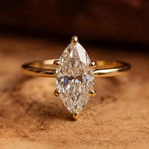 May include: A gold ring featuring a single marquise-cut clear gemstone held in place by six prongs. The band is smooth and polished, showcasing a simple design that highlights the elongated shape of the central stone and the metallic luster.