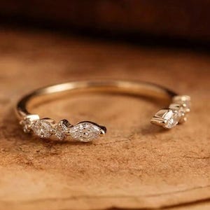 Op de afbeelding: Een open gouden ring met een reeks heldere, sprankelende stenen langs de band. Het ontwerp bevat een mix van ronde en marquise-geslepen stenen in een delicaat, asymmetrisch patroon dat een opening laat aan de bovenkant van de ring.
