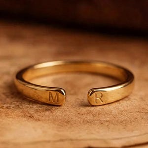 May include: A gold-toned open band ring featuring a smooth, polished finish. The two ends of the ring are rounded and each is engraved with a single capital letter, with the letter M on one side and the letter R on the other side of the opening.