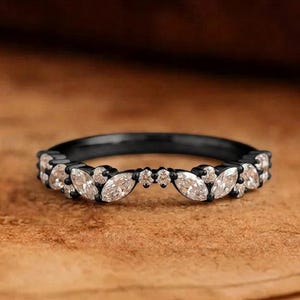 May include: A black metal ring featuring a curved band design adorned with a series of marquise-cut and round-cut clear stones. The stones are arranged in a symmetrical pattern along the front of the band, creating a delicate and detailed look.