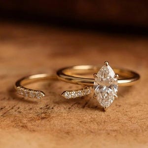 Könnte beinhalten: Ein Goldring-Set bestehend aus zwei Ringen. Ein Ring besitzt einen mittig platzierten Marquise-Schliff-Stein, der von sechs Krappen gehalten wird. Der zweite Ring ist ein offener Bandring mit einer Reihe kleiner Pavé-gefasster Steine an den Enden.