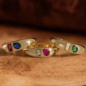 Puede incluir: Tres anillos de tono dorado, cada uno con una piedra rectangular de corte baguette y una piedra en forma de lágrima. El primer anillo tiene una piedra roja y una azul, el segundo una azul claro y una rosa, y el tercero una blanca y una verde.