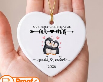 Puede incluir: Un adorno de cerámica blanco en forma de corazón con un cordón dorado. El texto dice "OUR FIRST CHRISTMAS AS MR & MRS" con un gráfico de flecha, dos pingüinos abrazados, corazones rosas, los nombres "sarah & robert" y el año "2026".