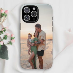 May include: A smartphone case featuring a printed photograph of a family of three standing on a beach at sunset. The image shows a person in a green dress holding a baby, while another person in a beige shirt stands beside them.