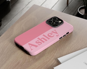 Puede incluir: Una funda para smartphone de color rosa en dos tonos con el nombre Ashley impreso en una fuente serif de color rosa oscuro en el centro. La funda incluye un módulo de cámara negro con tres lentes y botones laterales negros.