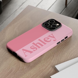 May include: A two-tone pink smartphone case featuring the name Ashley printed in a bold, dark pink serif font across the center. The case includes a black camera module cutout with three circular lenses and black side buttons for volume control.