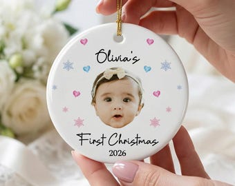 Puede incluir: Un adorno redondo de cerámica blanca con una fotografía central de un bebé con una diadema de lazo. El texto dice Olivia's First Christmas 2026, rodeado de pequeños corazones rosas, corazones azules y copos de nieve azules y rosas.