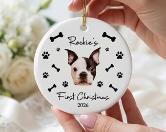 Puede incluir: Un adorno redondo de cerámica blanca con el retrato central de un perro blanco y negro. El texto dice Rockie's First Christmas 2026. El diseño incluye huellas de patas y huesos negros dispersos alrededor del perro, colgado de un cordón dorado.