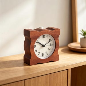 Può includere: Una sveglia quadrata in legno con finitura marrone scuro e una cornice ondulata e sagomata. Il quadrante è bianco con numeri arabi neri, lancette nere per ore e minuti e una sottile lancetta dei secondi nera. Un piccolo pulsante bianco è in alto.