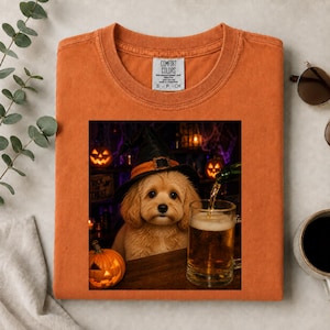 Puede incluir: Una camiseta naranja con el gráfico de un cachorro de color claro que lleva un sombrero de bruja negro, sentado junto a una jarra de cerveza y una pequeña calabaza. El fondo muestra una escena oscura y festiva de Halloween con calabazas.