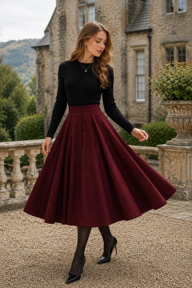 May include: A long-sleeved black ribbed jumper paired with a high-waisted, pleated burgundy midi skirt. The skirt features a vertical row of matching buttons down the centre front, creating a structured, flared silhouette that falls below the knees.
