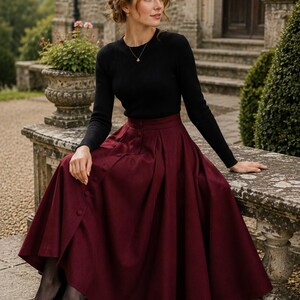 May include: A long, deep burgundy skirt featuring a high waist, a row of matching buttons down the front, and deep pleats, paired with a black long-sleeved ribbed jumper, sheer black tights, and black pointed-toe high-heeled pumps.