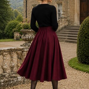 May include: A long, pleated, deep burgundy skirt with a high waist and a flared silhouette, paired with a black long-sleeved top, sheer black tights, and black high-heeled pumps. The skirt features a smooth, solid-coloured fabric and a fitted waistband.