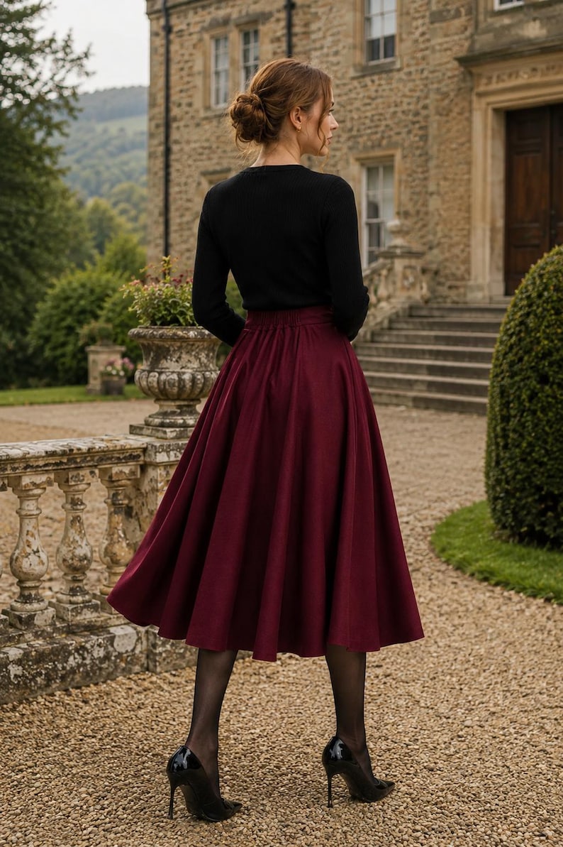 May include: A long, pleated, deep burgundy skirt with a high waist and a flared silhouette, paired with a black long-sleeved top, sheer black tights, and black high-heeled pumps. The skirt features a smooth, solid-coloured fabric and a fitted waistband.