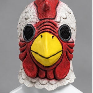May include: A full-head chicken mask featuring a textured white feathered surface, a bright yellow beak, and a prominent red comb and wattle. The mask includes two circular black mesh eye openings positioned on the sides of the head.