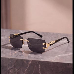 Puede incluir: Gafas de sol sin montura con lentes rectangulares de color gris oscuro, puente y bisagras de metal dorado y patillas negras. Las bisagras están adornadas con una figura de pantera dorada, aportando un acento metálico al diseño.