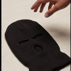 May include: A solid black knitted balaclava featuring a ribbed texture throughout the fabric. The headgear includes three distinct openings: two elongated eye holes positioned side by side and one smaller, centered mouth opening below them.