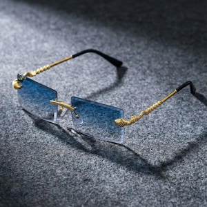 Puede incluir: Gafas de sol rectangulares sin montura con lentes de degradado azul. Las patillas de metal dorado presentan un motivo de serpiente texturizado en las bisagras y terminan en puntas de plástico negro para un ajuste seguro.