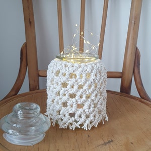 May include: A white crocheted jar cover with a glass jar and string lights inside, sitting on a wooden chair.