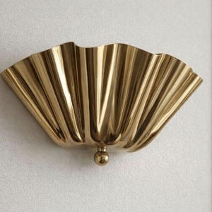 May include: A wall-mounted light fitting with a polished gold finish. The lamp features a wide, fan-shaped shade with deep, vertical pleats that create a scalloped edge. A small, spherical gold knob is positioned at the base of the pleated shade.