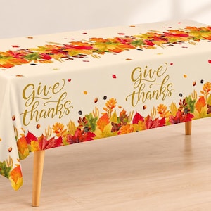 May include: A rectangular cream-colored tablecloth featuring a repeating pattern of colorful autumn leaves in shades of red, orange, yellow, and green, with the gold cursive text "Give thanks" printed across the fabric surface.