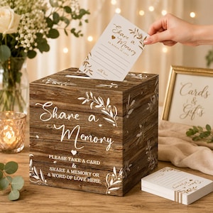 May include: A square wooden box with a dark brown wood grain finish, featuring white text that reads Share a Memory and Please take a card & share a memory or a word of love here, decorated with white olive branch illustrations.
