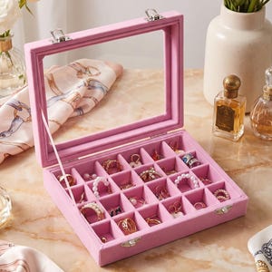 May include: A rectangular pink velvet jewelry organizer box with a clear glass lid and silver metal latches. The interior features twenty-four individual square compartments, each containing various gold and silver rings, earrings, and pearl jewelry.