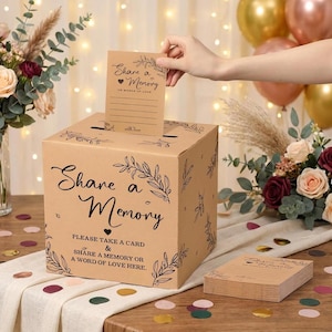 May include: A square cardboard memory box with a light brown finish, featuring black text that reads Share a Memory and Please take a card & share a memory or a word of love here, decorated with black line art of leaves and a small heart icon.