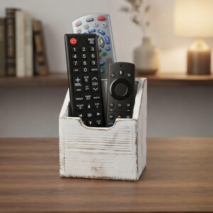 May include: A white distressed wooden remote control holder with a rectangular base and an open top. The holder contains three remote controls: one black remote with a number pad, one silver remote, and one black streaming device remote.