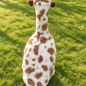 May include: A plush toy shaped like a cow with a long neck, featuring a white body covered in brown patches. The toy has two tan horns at the top of its head and small brown ears on the sides. The texture of the fabric appears soft and fuzzy.