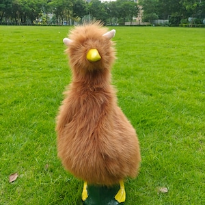 May include: A plush toy shaped like a bird with long, shaggy brown fur, two small light-colored horns on top of its head, a bright yellow beak, and two yellow webbed feet standing on a dark green base, creating a whimsical creature design.