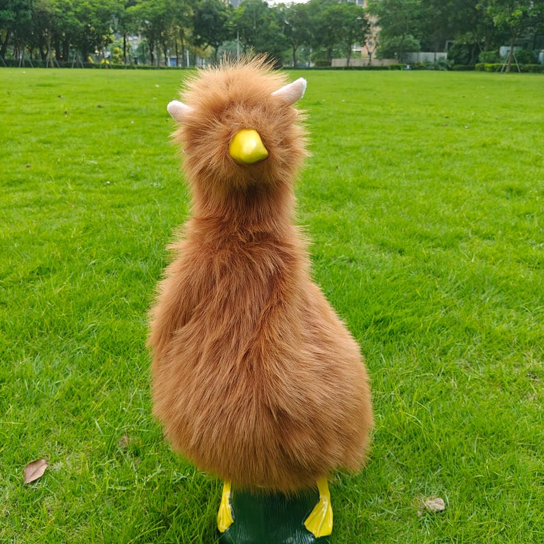 May include: A plush toy shaped like a bird with long, shaggy brown fur, two small light-colored horns on top of its head, a bright yellow beak, and two yellow webbed feet standing on a dark green base, creating a whimsical creature design.