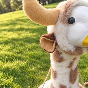 May include: A plush giraffe head wall mount featuring soft white and brown spotted fur, a smooth white face, a yellow nose, and a single prominent tan felt horn. The giraffe has a small brown-lined ear and is attached to a light wooden base.