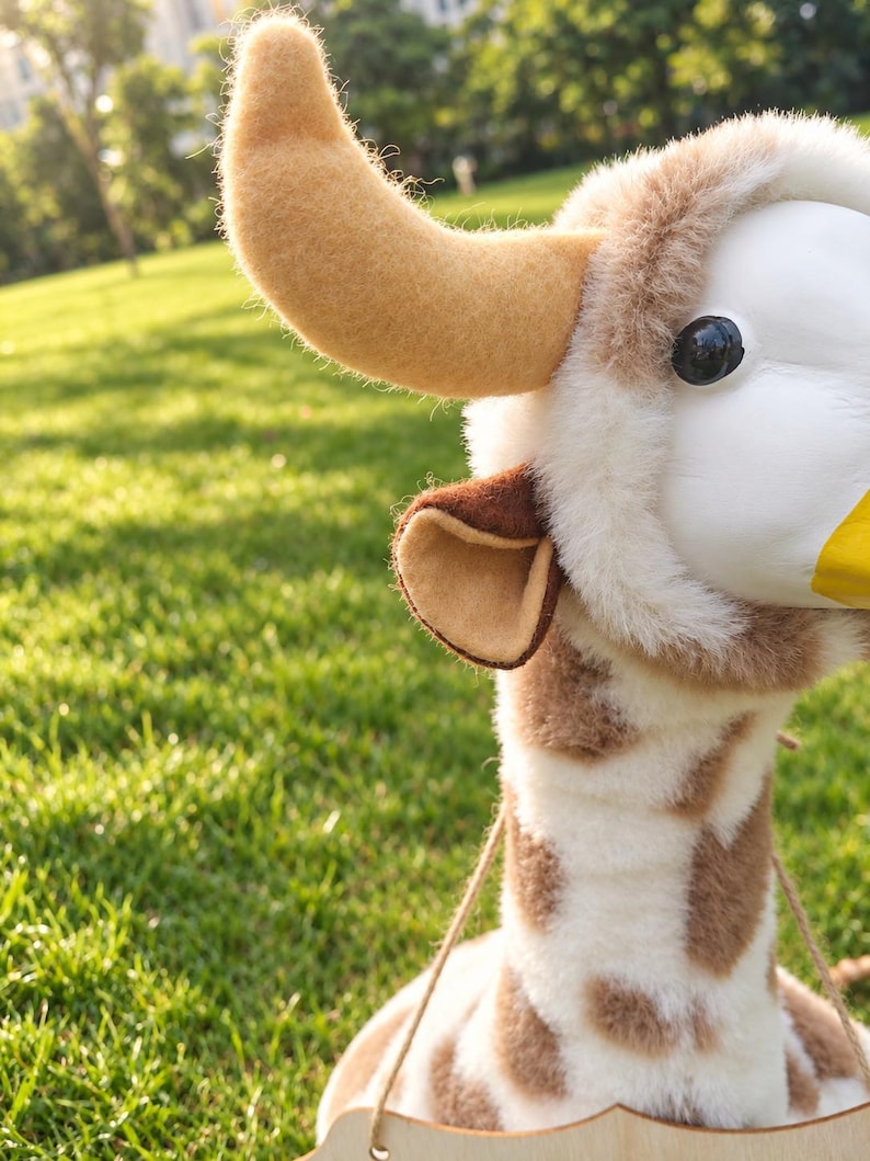 May include: A plush giraffe head wall mount featuring soft white and brown spotted fur, a smooth white face, a yellow nose, and a single prominent tan felt horn. The giraffe has a small brown-lined ear and is attached to a light wooden base.