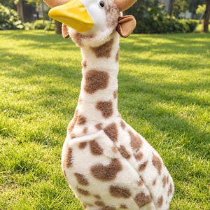 May include: A plush goose toy featuring a white and brown giraffe-patterned body, a yellow beak, and two tan felt horns attached to the head. The figure has a long neck, soft textured fur, and small dark eyes, standing upright on yellow feet.
