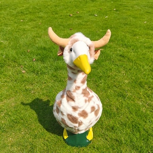 May include: A plush goose figurine featuring a white and brown spotted pattern, a bright yellow beak, and two tan-colored horns protruding from the top of its head. The figure has a smooth white face and stands on a solid dark green base.