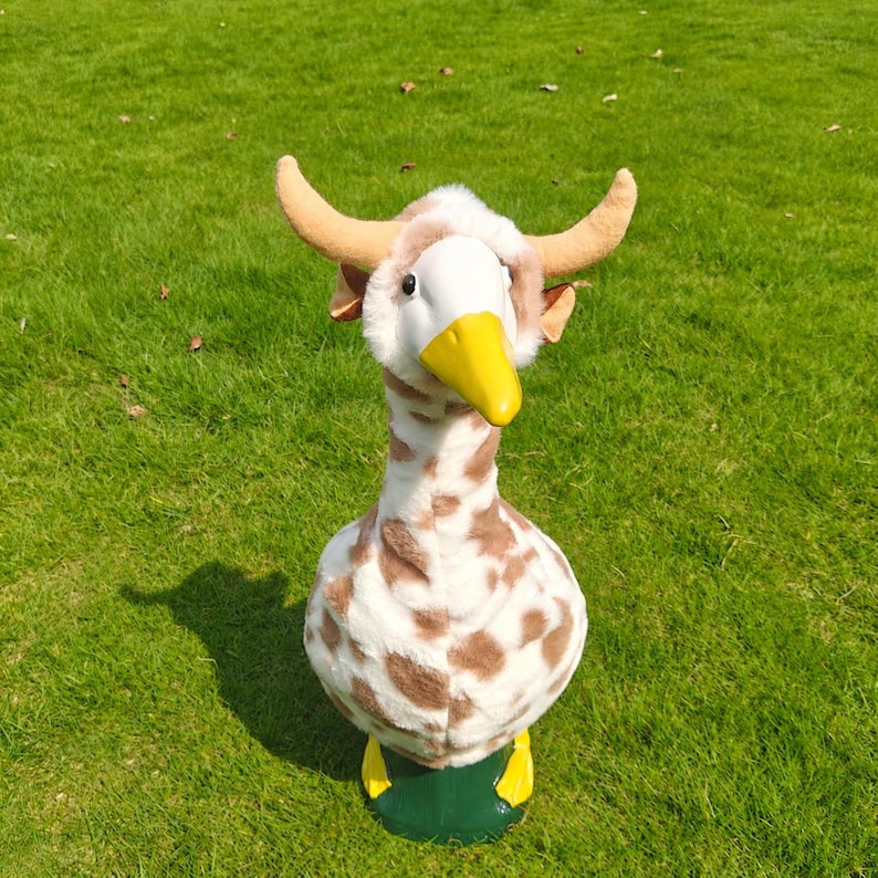 May include: A plush goose figurine featuring a white and brown spotted pattern, a bright yellow beak, and two tan-colored horns protruding from the top of its head. The figure has a smooth white face and stands on a solid dark green base.
