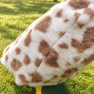May include: A plush toy featuring a soft, white and brown spotted faux fur texture resembling a giraffe pattern. The toy has a rounded body shape and is supported by a solid, dark green base with a bright yellow, webbed foot visible at the bottom.