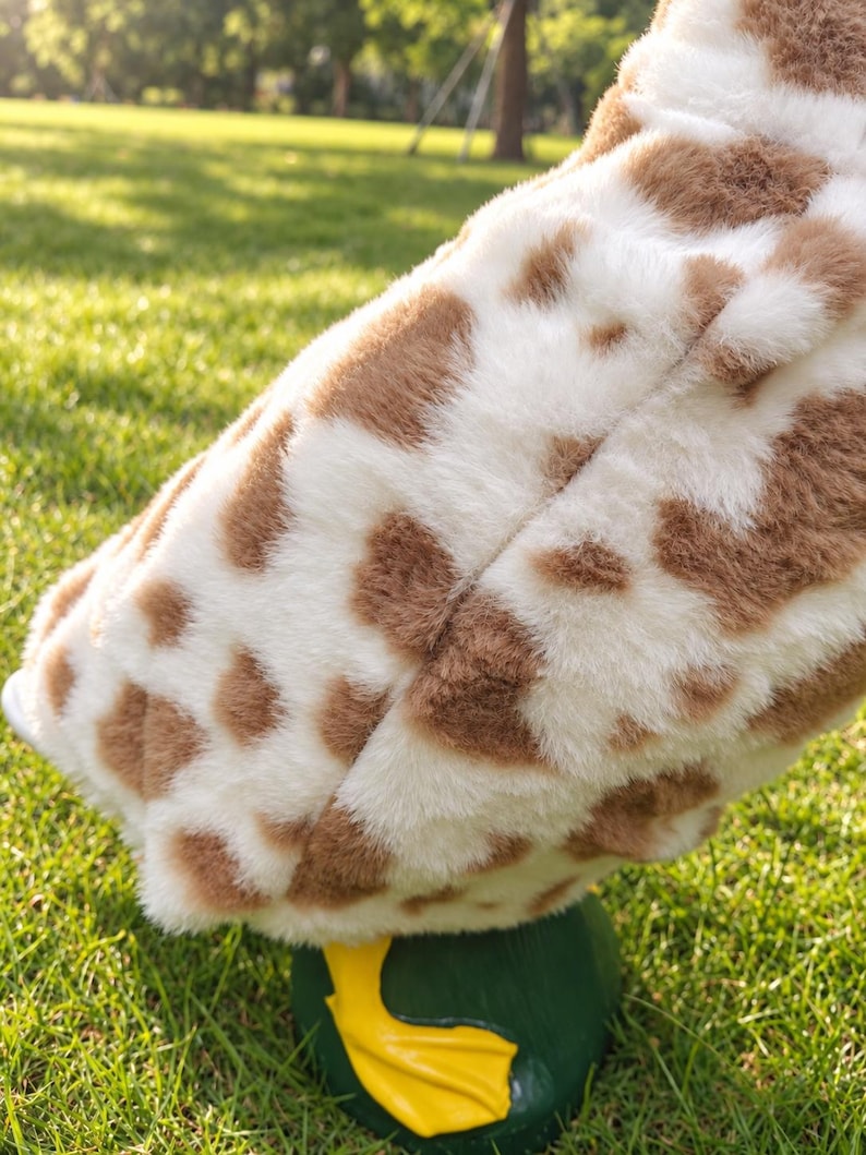 May include: A plush toy featuring a soft, white and brown spotted faux fur texture resembling a giraffe pattern. The toy has a rounded body shape and is supported by a solid, dark green base with a bright yellow, webbed foot visible at the bottom.