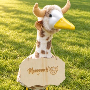 May include: A plush toy featuring a white and brown spotted giraffe-like body with a smooth white face, a yellow beak, and two tan horns. A light-colored wooden sign hangs from its neck, engraved with the text Moooooo!! and a cow illustration.