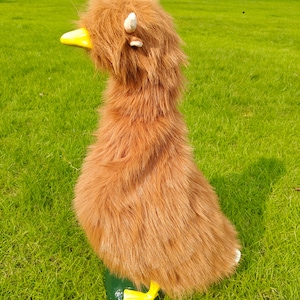 May include: A plush stuffed animal shaped like a duck, covered in long, shaggy, light brown faux fur. The toy features a bright yellow beak, small white tufts representing ears or feathers on the head, and yellow feet at the base.