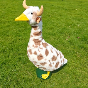 May include: A plush goose toy featuring a white and brown spotted pattern, a bright yellow beak, and two small tan horns protruding from the top of its head. The stuffed animal has a long neck and stands on two yellow webbed feet.