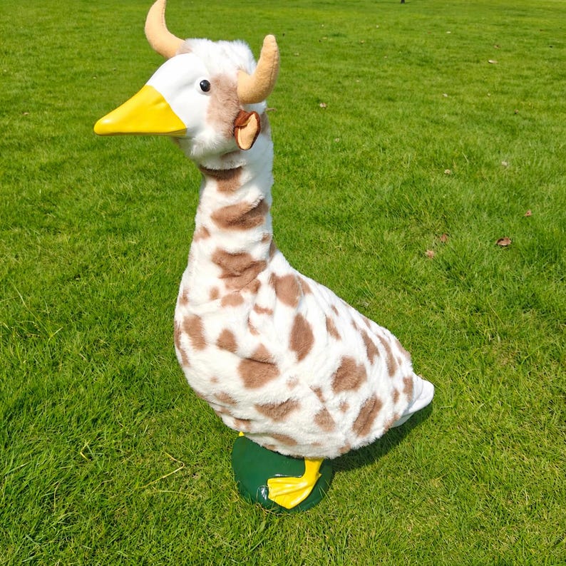 May include: A plush goose toy featuring a white and brown spotted pattern, a bright yellow beak, and two small tan horns protruding from the top of its head. The stuffed animal has a long neck and stands on two yellow webbed feet.