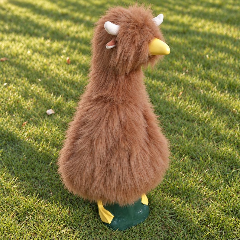 May include: A plush duck figurine covered in long, shaggy brown faux fur. The duck features a smooth, solid yellow beak, two small white horns protruding from the top of its head, and bright yellow webbed feet attached to a dark green base.