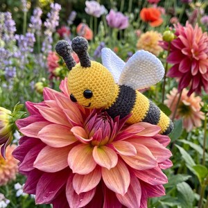 Könnte beinhalten: Ein gehäkeltes Bienen-Spielzeug mit einem gelb-schwarz gestreiften Körper, zwei dunkelgrauen Fühlern und weißen Flügeln. Die Biene hat zwei kleine schwarze Kunststoffaugen und ein aufgesticktes Lächeln auf einer großen, pink-orangen Dahlie.